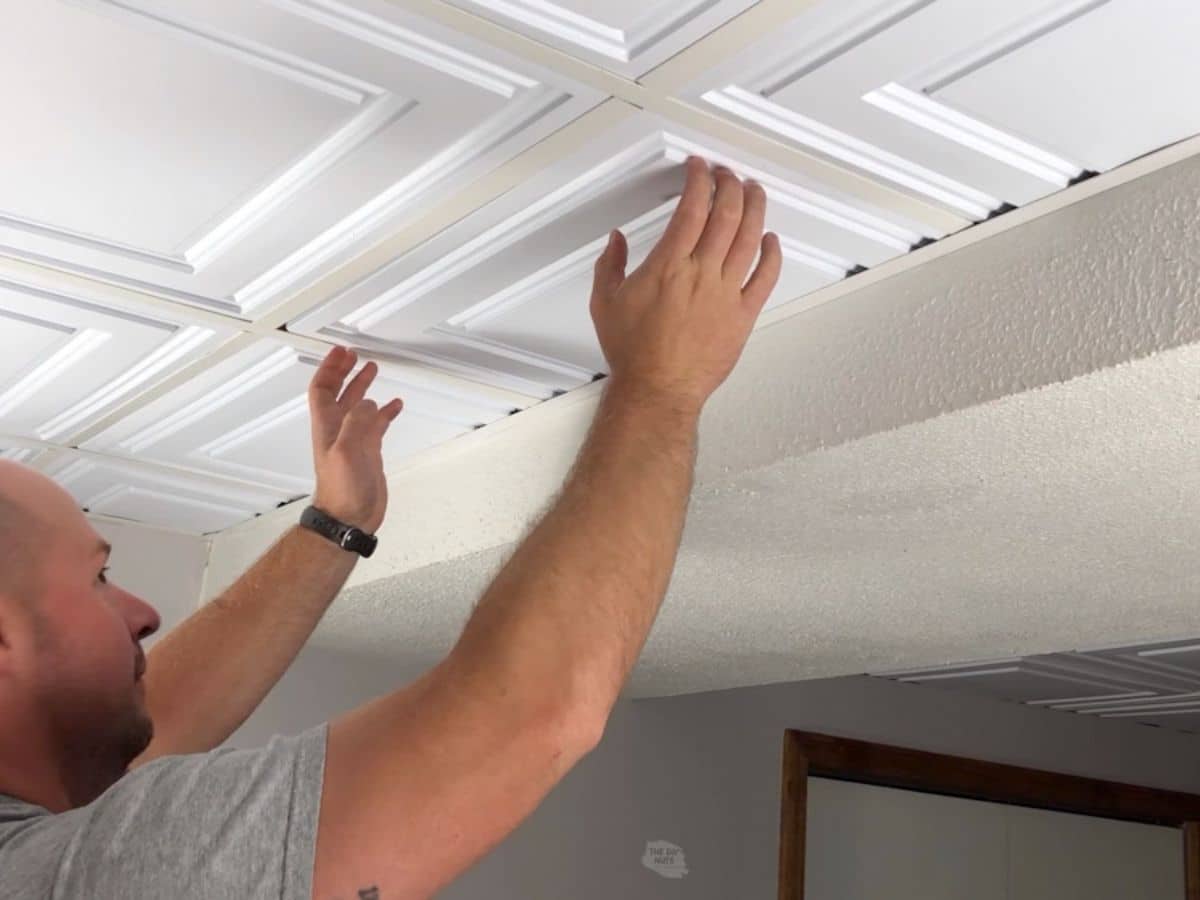 man adding white drop ceiling tiles to basement ceiling.